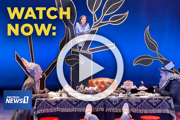 Watch Now. A play button overlays a scene from the Mad Tea Party in Alice in Wonderland. Alice gazes from a black tree outlined in gold as the March Hare in a knitted beanie and the Mad Hatter in a top hat laugh over a table of cakes and tea.